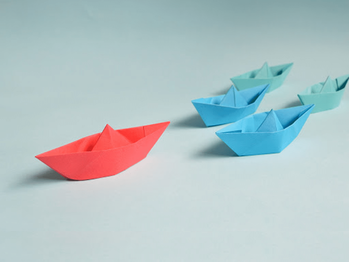 A red paper boat, exemplifying strong leadership, leads a group of blue paper boats against a light blue background, symbolizing guidance and direction.