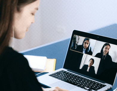 A woman participates in a video conference on a laptop, with three other people visible on the screen. She is sitting at a desk with a stack of notebooks beside her, engaging in a professional discussion, sharing her experience and insights about our practices.