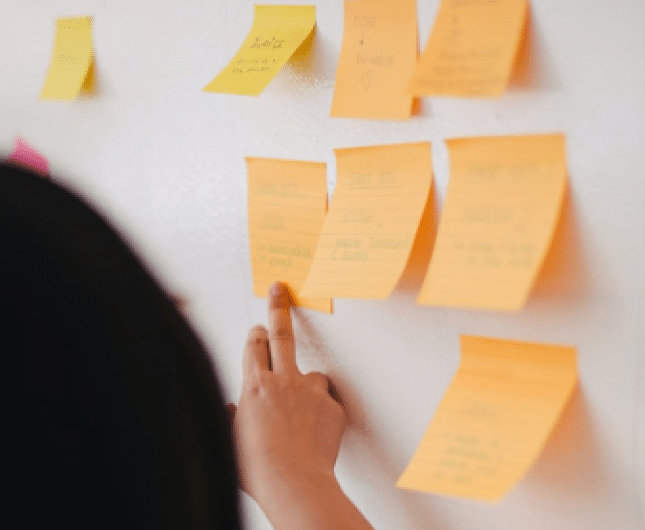 A person with long dark hair is seen from behind, placing an orange sticky note on a white wall filled with other sticky notes. The notes, likely detailing sales strategies, have handwritten text on them. The setting suggests brainstorming or planning practices.