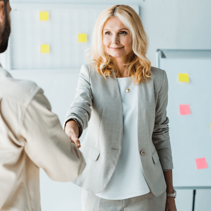 A woman with blonde hair, dressed in a light grey suit, is smiling and shaking hands with a person whose back is turned to the camera. They are in a creative agency office setting with colorful sticky notes on whiteboards in the background, suggesting a lively recruiting meeting.