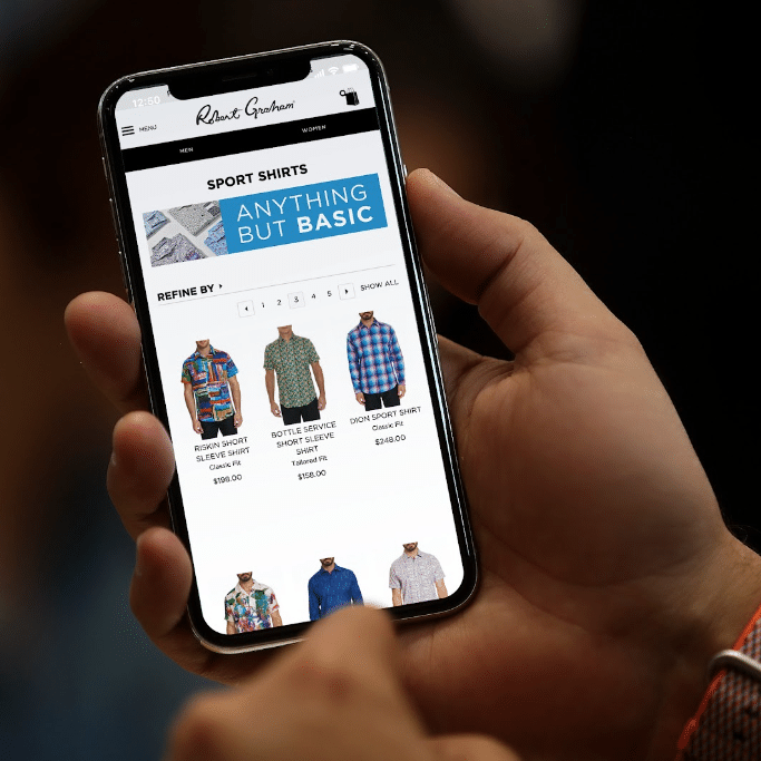 A VP holds a smartphone displaying an online store page for Robert Graham sport shirts. Various shirt styles and prices are visible on the screen. The executive's thumb and forefinger are positioned near the bottom, as if scrolling or selecting an item in their ecommerce app.