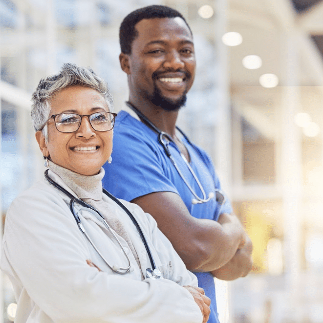 Two healthcare professionals are standing and smiling. The woman on the left has short gray hair, glasses, and is wearing a white lab coat with a stethoscope. The man on the right has short black hair, is wearing blue scrubs, and also has a stethoscope. Perfect for recruiting or marketing materials in healthcare.