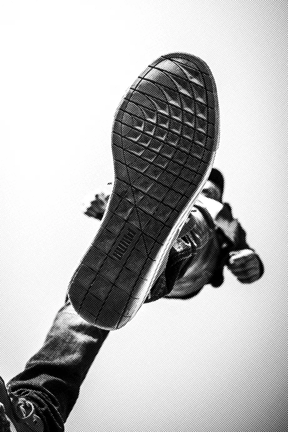 A black-and-white image of a close-up of the bottom tread of a branded sneaker. The shoe is in focus and appears to be worn by someone who is either jumping or stepping forward, with the sky in the background. The tread pattern is clearly visible.