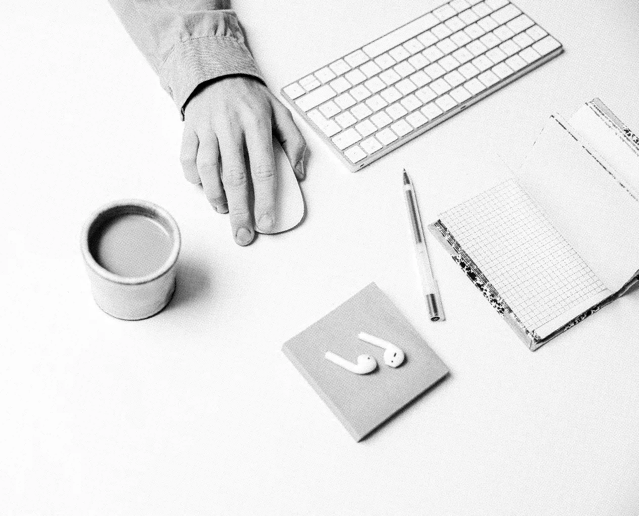 A monochrome image displays a minimalist workspace with a person's hand on a computer mouse, a wireless keyboard, a pen, a notebook, wireless earbuds on a notepad, and a coffee cup. Our approach suggests this clean and organized environment fosters productivity.