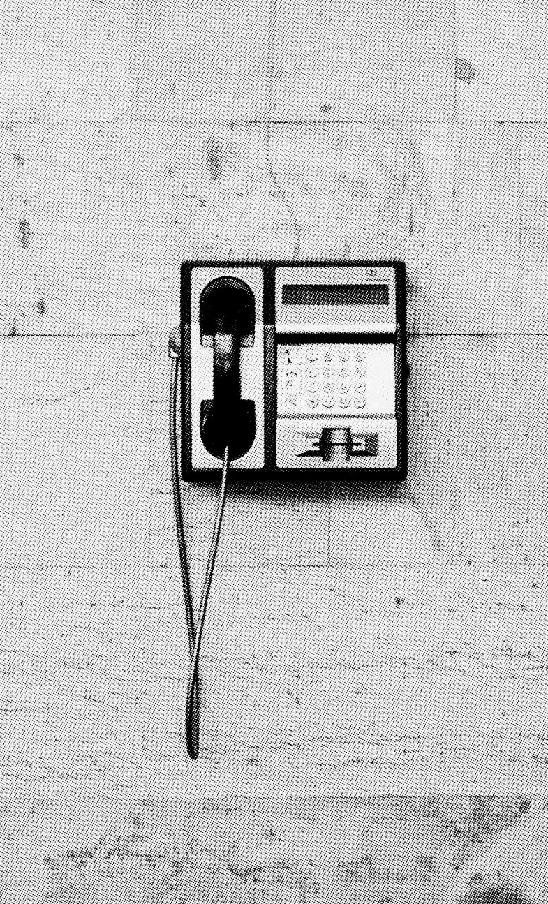 Black and white image of a vintage payphone mounted on a textured wall. The payphone has a keypad, coin slot, and a corded handset hanging to the left side. The wall has a patchy appearance with various shades of gray, inviting us to contact the past in subtle hues.