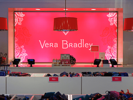 A retail display featuring a bright pink backdrop with "Vera Bradley" written in white demonstrates effective marketing practices. Red light fixtures hang from the ceiling, illuminating the display. Tables in the foreground showcase various products, with discount signs visible.