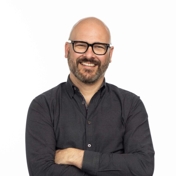 A bald man with glasses, a beard, and a mustache smiles while crossing his arms. He is wearing a button-up long-sleeve dark shirt against a plain white background, reminiscent of Freddie Laker's iconic style.